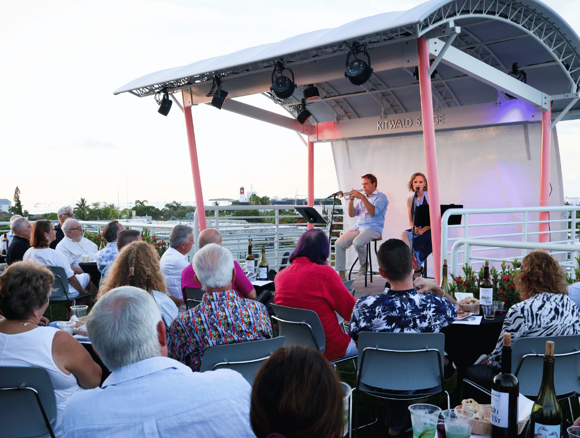 A trumpeter and a singer perform on the canopied Kitwald Stage at Hugh's View while an audience watches from cocktail tables, with the water visible beyond the rooftop railing.