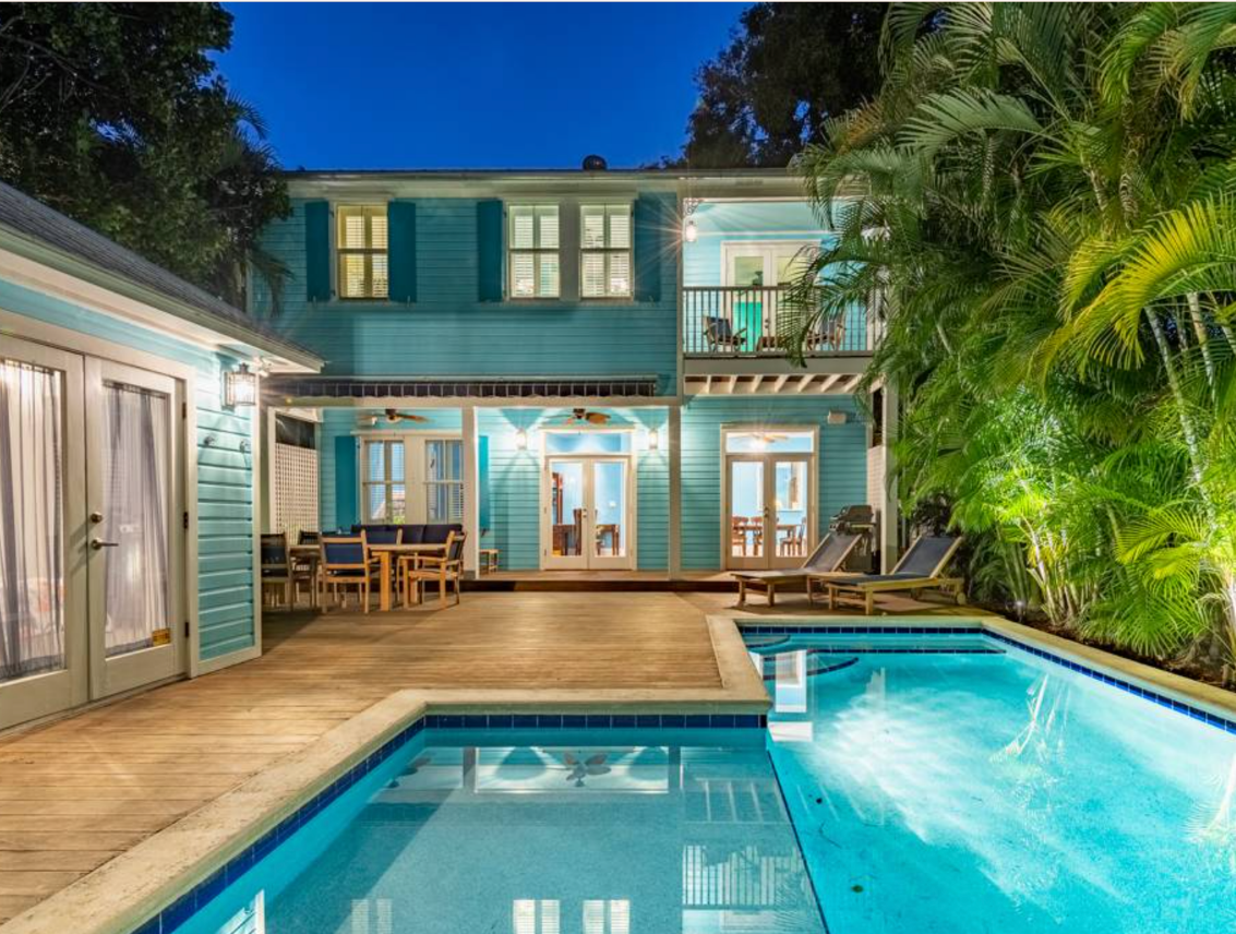 A turquoise two-story house at dusk with a lit swimming pool in the foreground, wooden deck, covered porch with dining table, and palms along the right side.