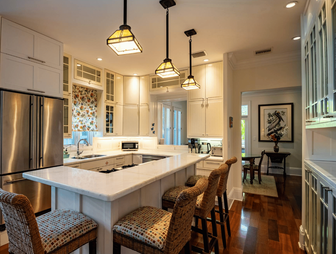 A warmly lit kitchen with cream cabinetry, a white marble peninsula counter, wicker barstools, and three glass pendant lights, opening onto a dining area with dark wood floors.