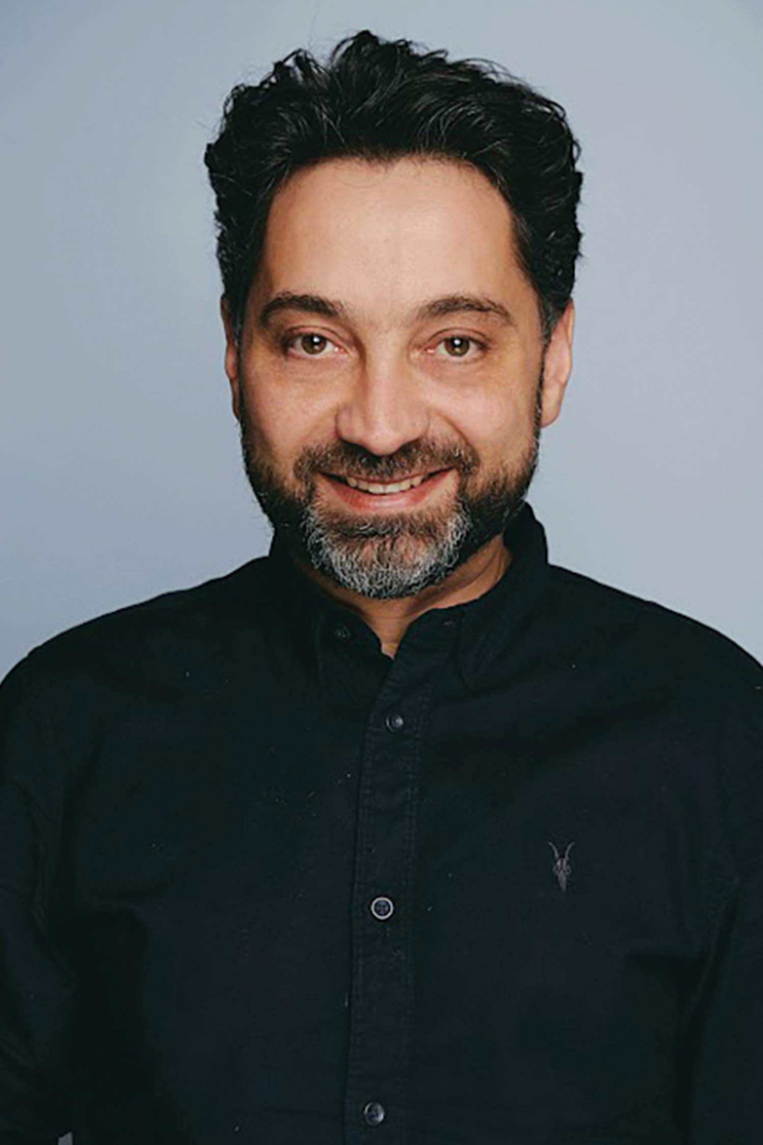 Head-and-shoulders portrait of a smiling man with dark wavy hair and a salt-and-pepper beard, wearing a black button-down shirt against a light gray background.