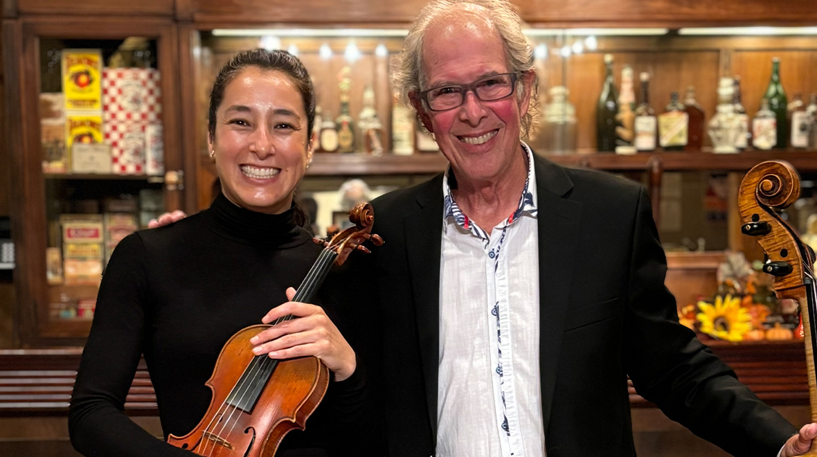 Two smiling musicians pose together, one holding a violin and the other standing beside a cello.