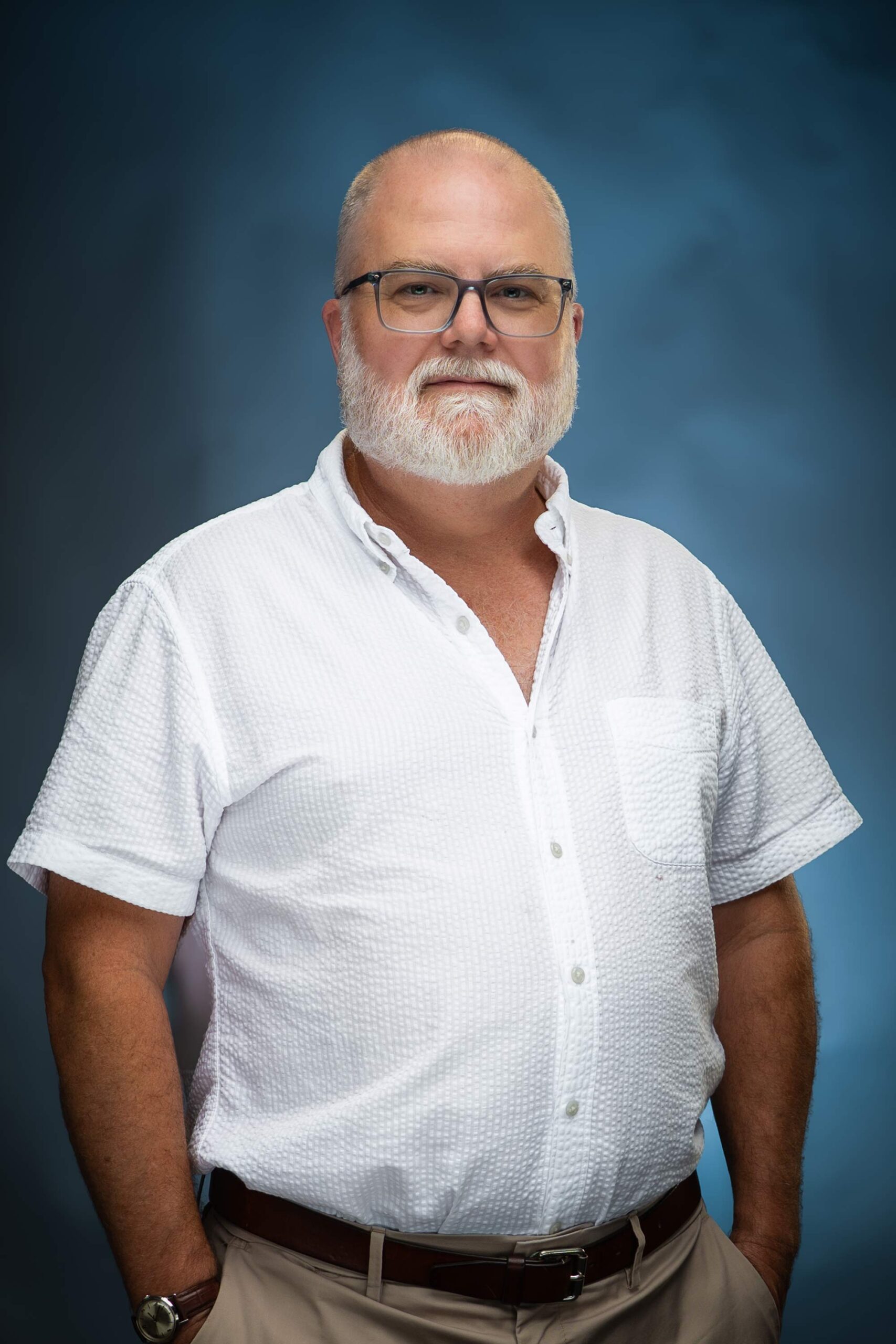Headshot of Jed in glasses and a white button-down shirt against a blue studio background.
