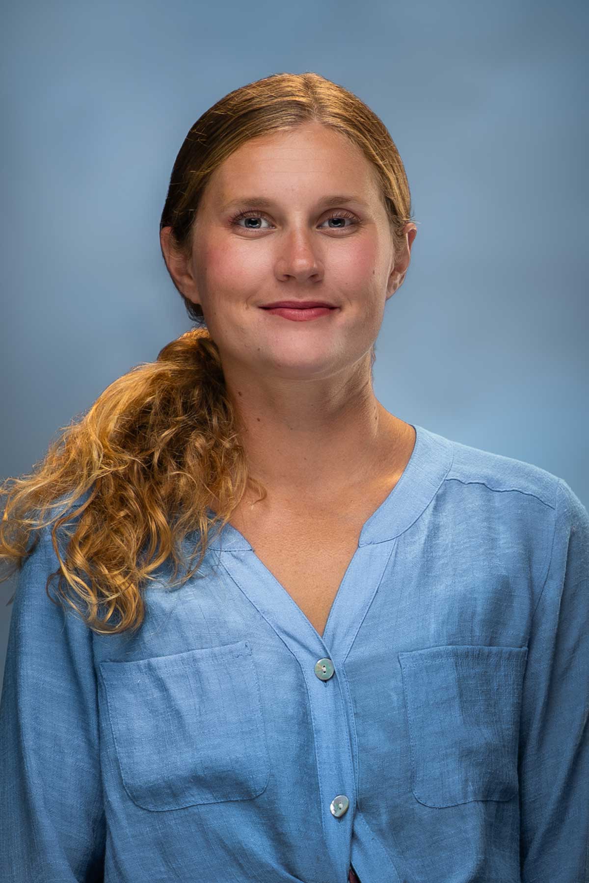 Headshot of a young woman with curly strawberry blonde hair in a side ponytail wearing a blue blouse.