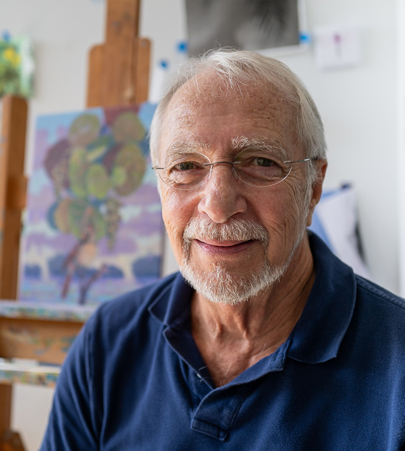 Jim Salem smiling in his studio, with a colorful painting on an easel behind him.