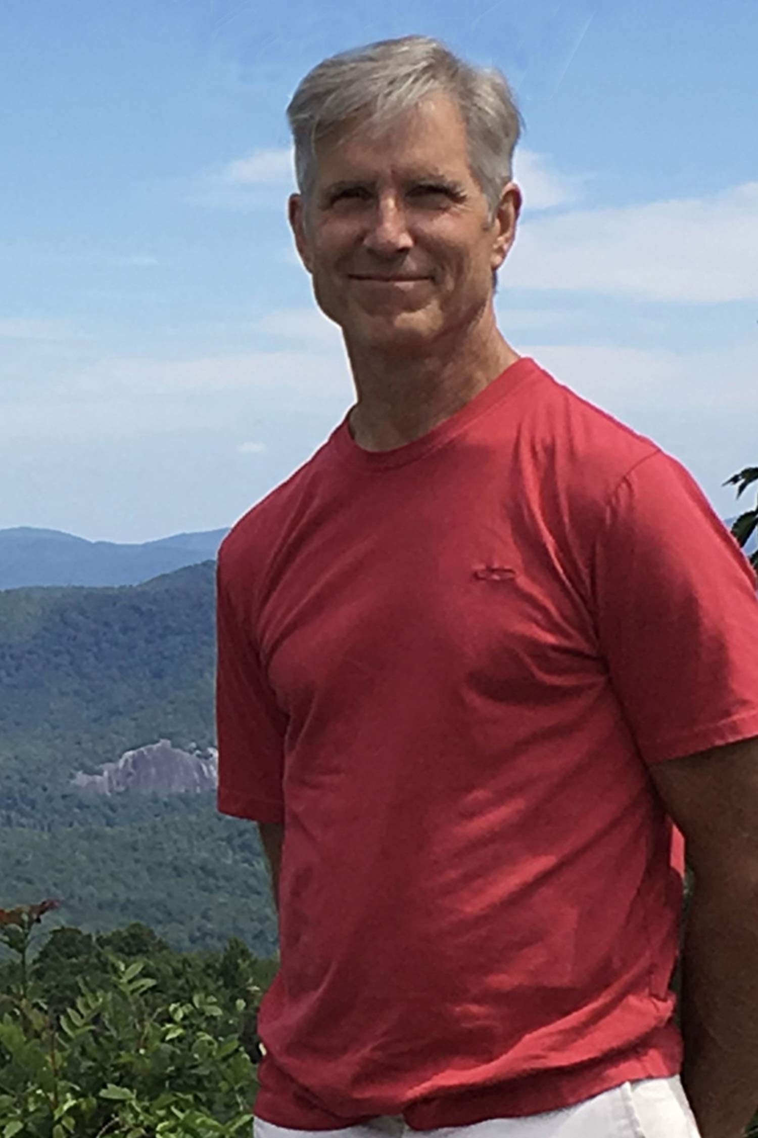 Jim Ostlund stands outdoors in a red shirt with forested mountains and a blue sky in the background.