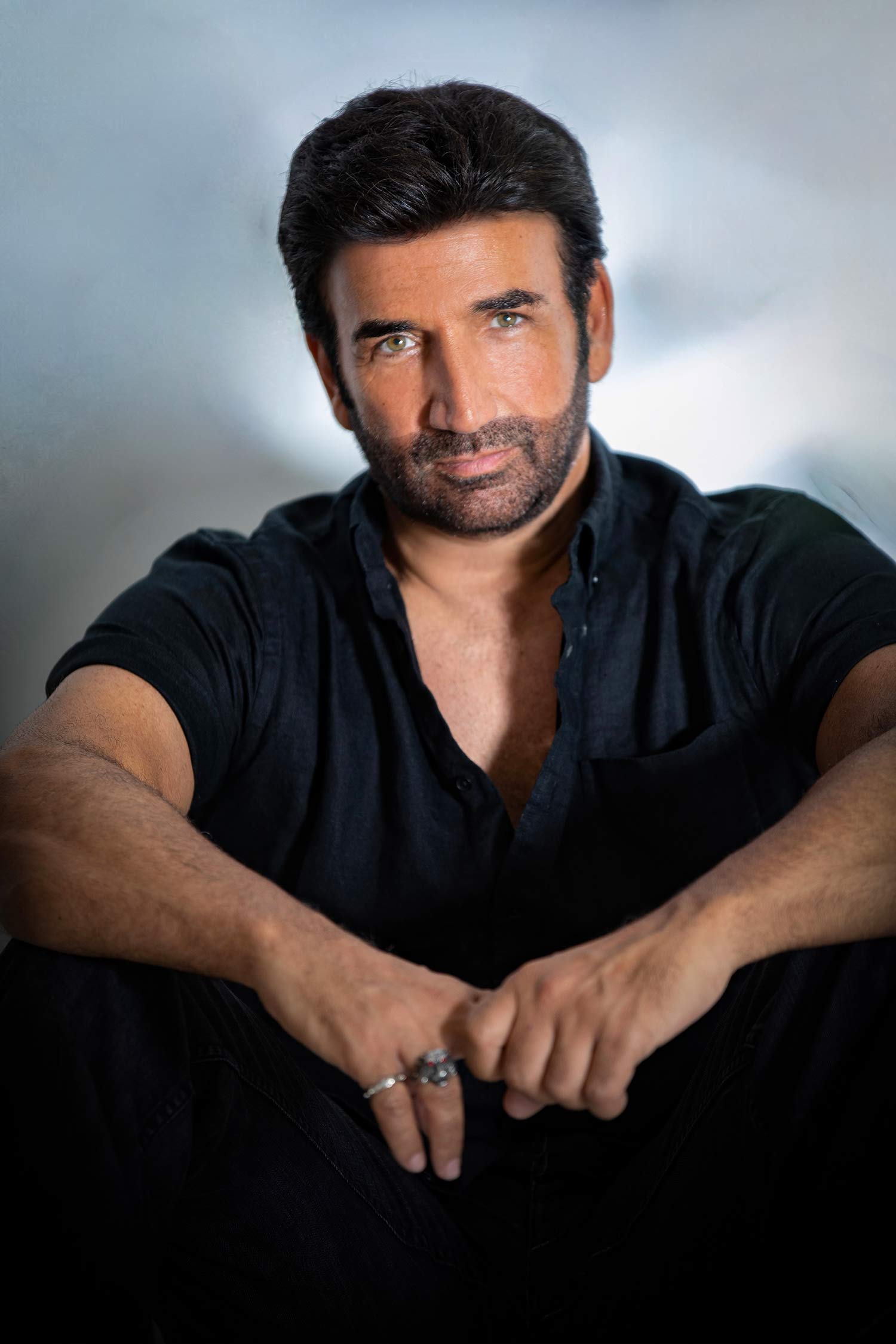 Portrait of a seated man with dark hair and a short beard, wearing a black shirt and rings, with his arms resting across his knees against a softly blurred background.