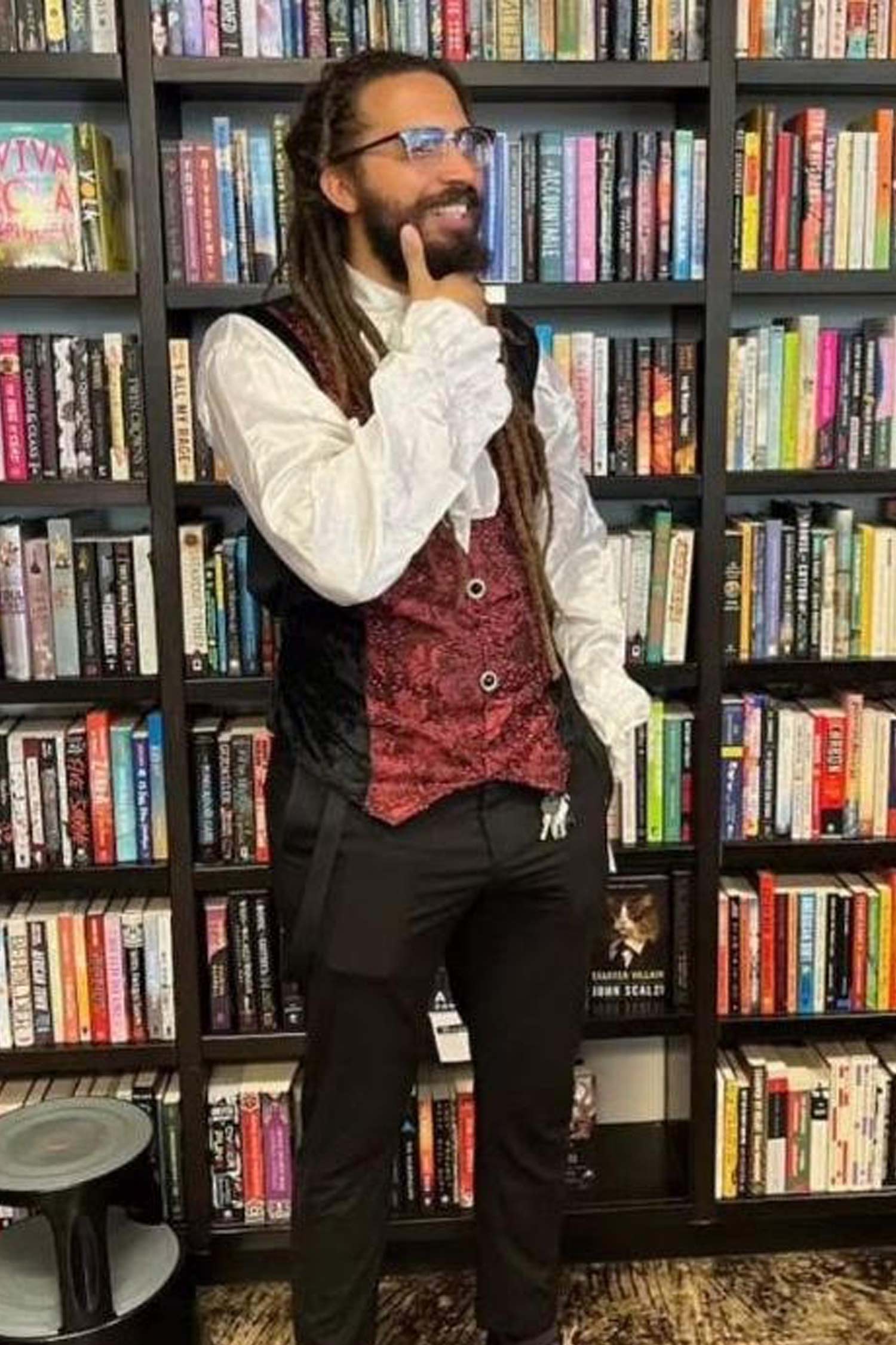 Smiling man with long locs, glasses and a beard wearing a red vest and white shirt, standing in front of bookstore shelves.