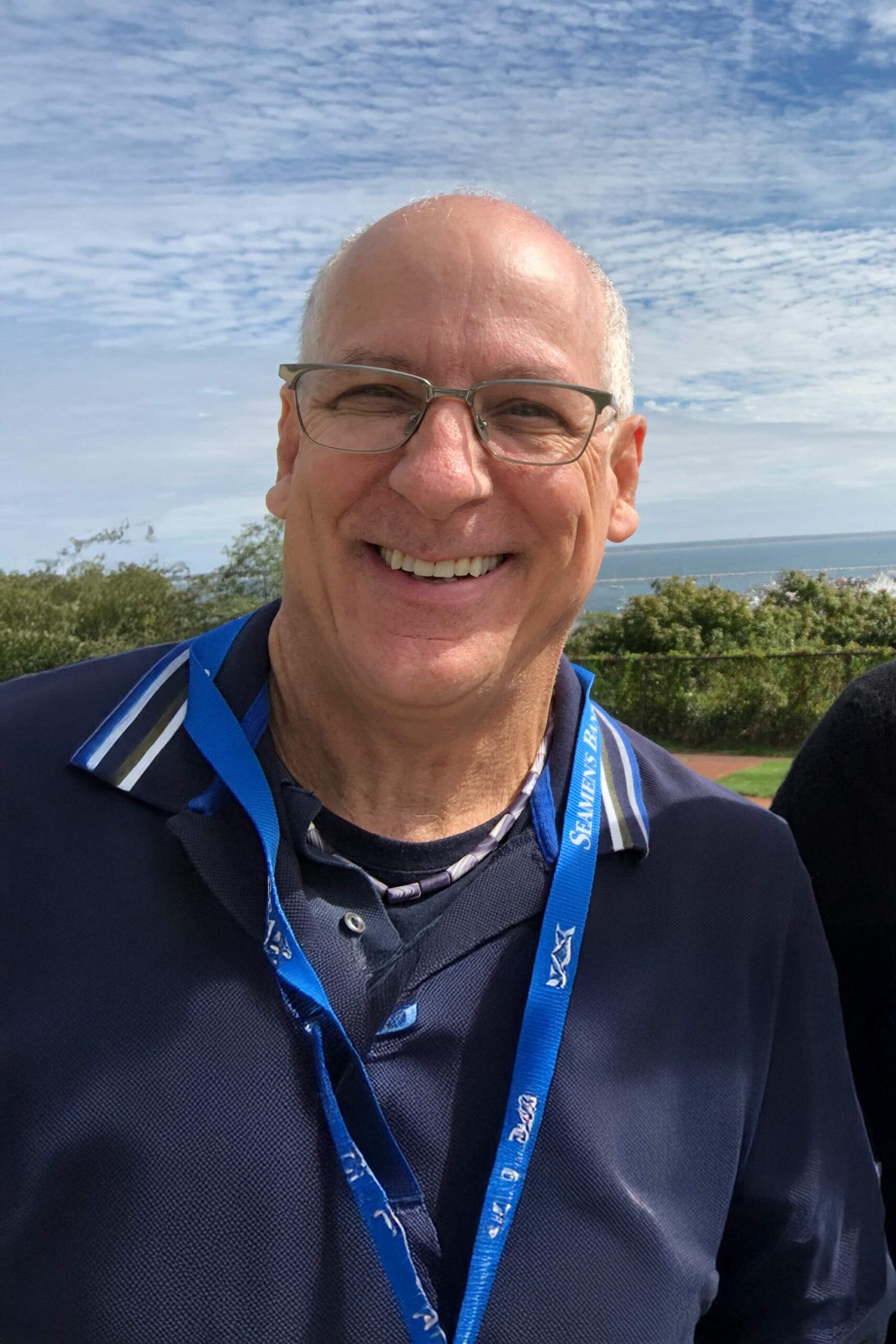 Outdoor headshot of John smiling in glasses, with blue sky and water in the background.