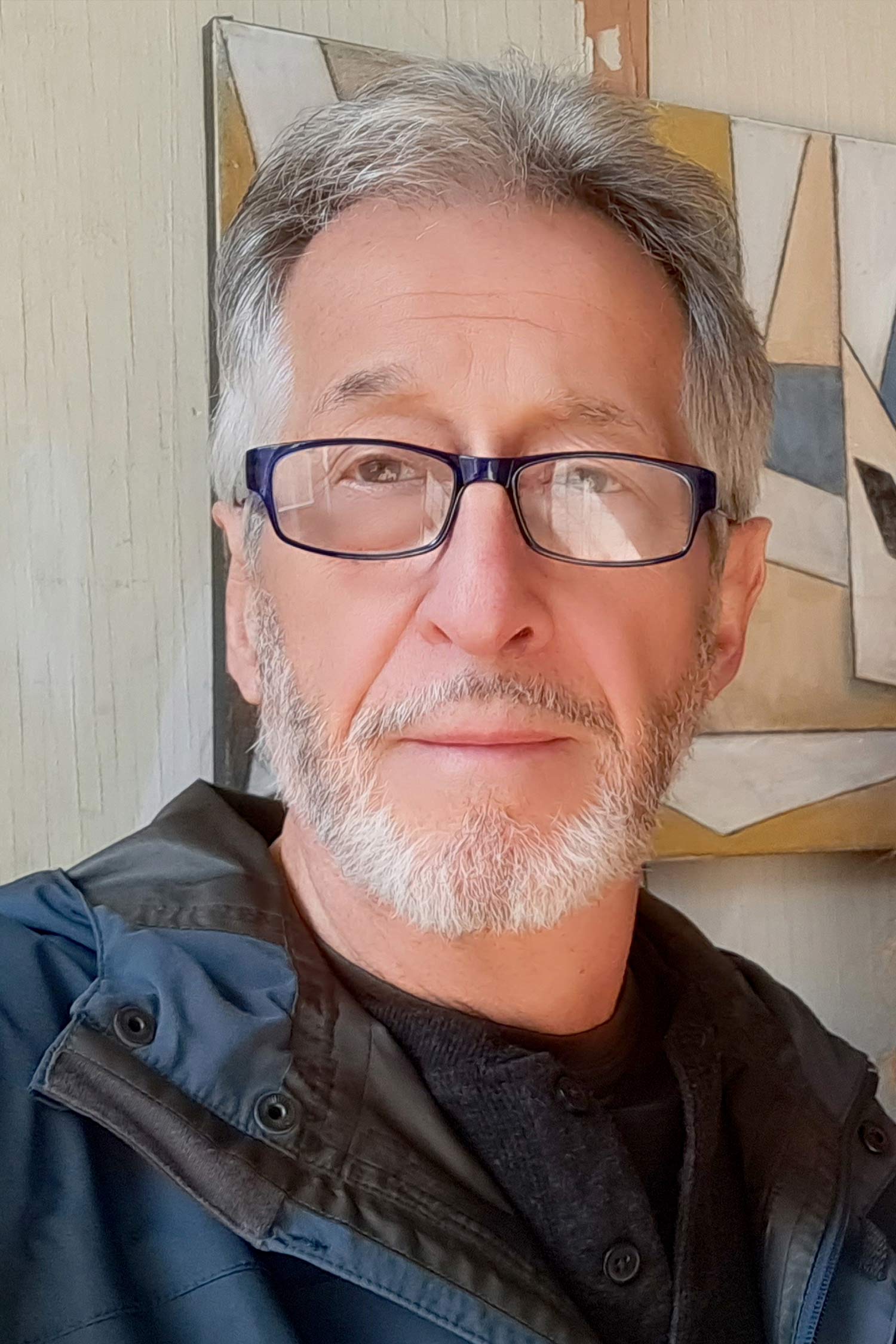 Close-up portrait of a man with short gray hair, a trimmed gray beard and rectangular glasses, with abstract artwork in the background.