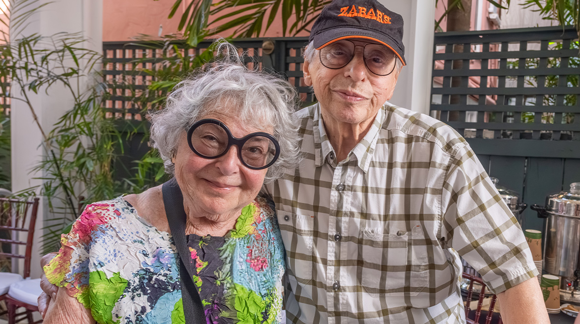 Older couple with gray hair and glasses smiling together at an outdoor gathering surrounded by tropical plants.