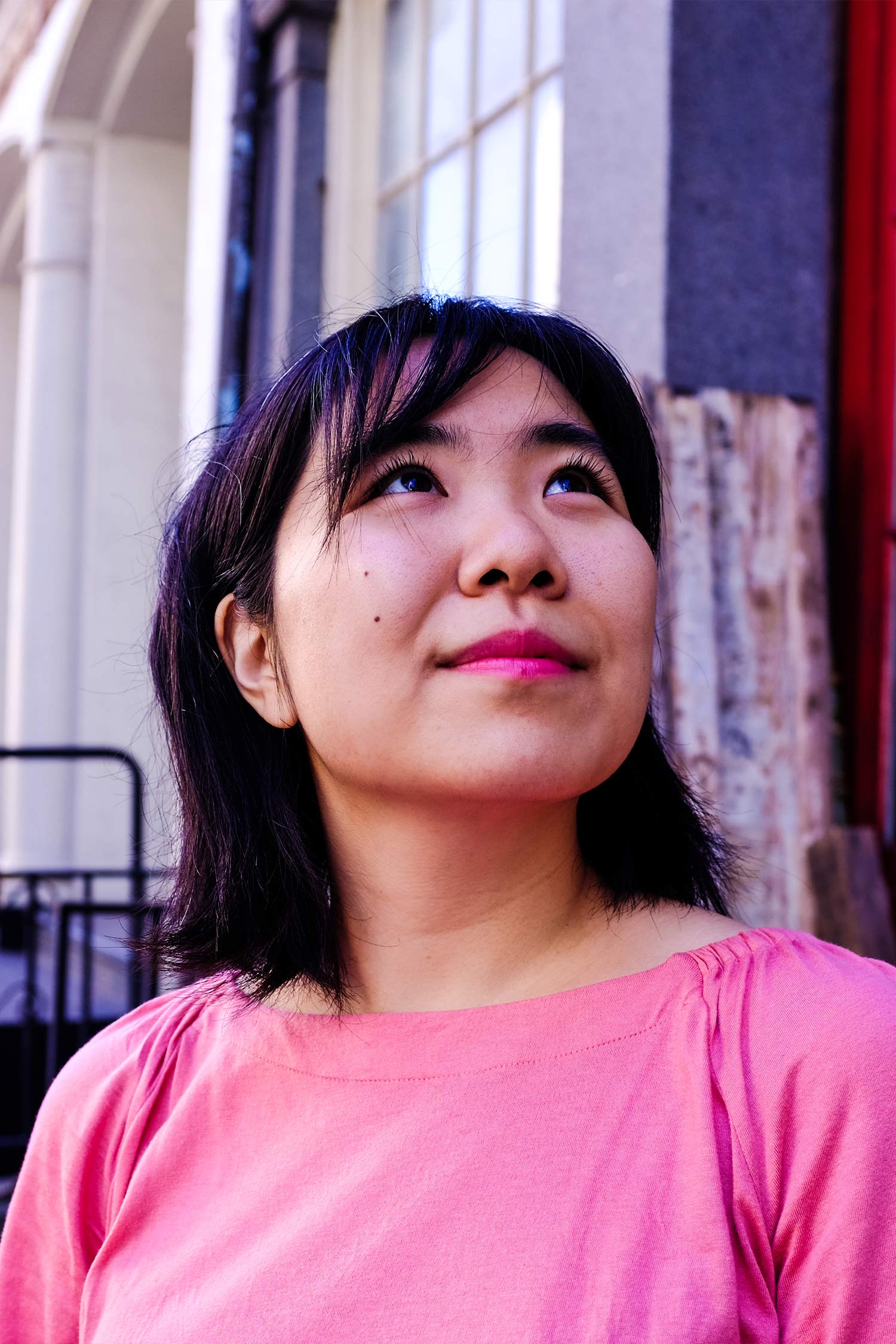 Close-up portrait of a woman with shoulder-length black hair and bangs, wearing a bright pink top and looking upward.