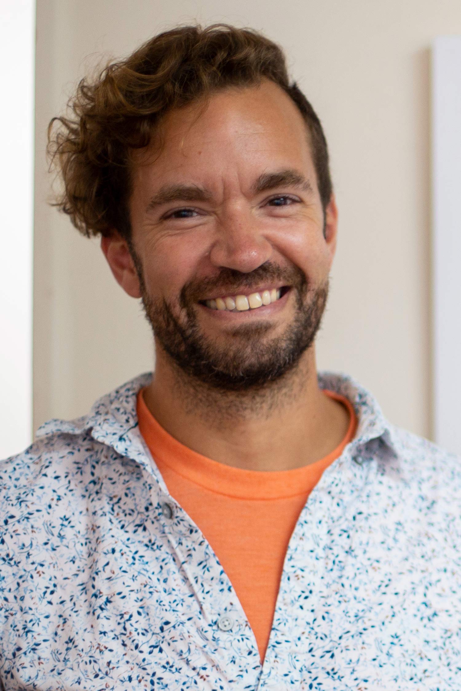 Smiling man with curly brown hair and a short beard, wearing an orange shirt beneath a light patterned button-down.