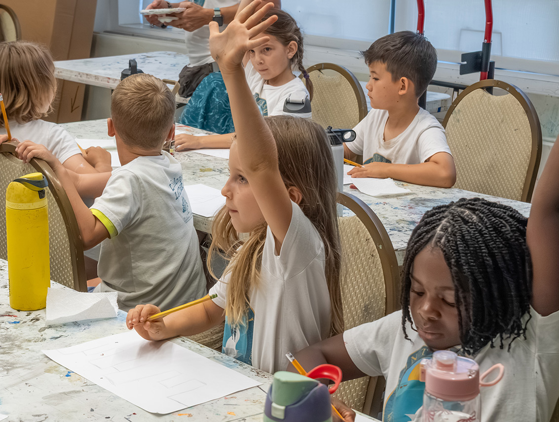 Children work on drawings around classroom tables as one student raises her hand during an art class.