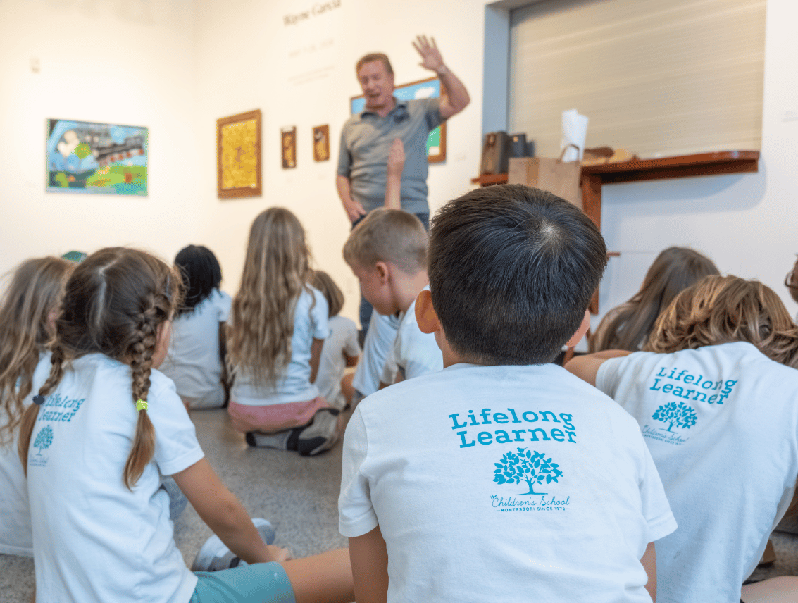 A group of schoolchildren sits on a gallery floor listening to an artist who gestures as he speaks, with framed artwork on the walls behind him.