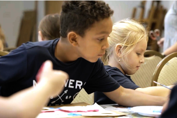 Two children in navy school shirts lean over a table, absorbed in an art project with colored paper and drawing materials in front of them