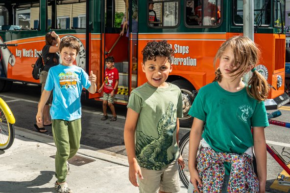 Three children stand smiling on a sunny sidewalk in front of an orange and green Old Town Trolley, one giving a thumbs-up.