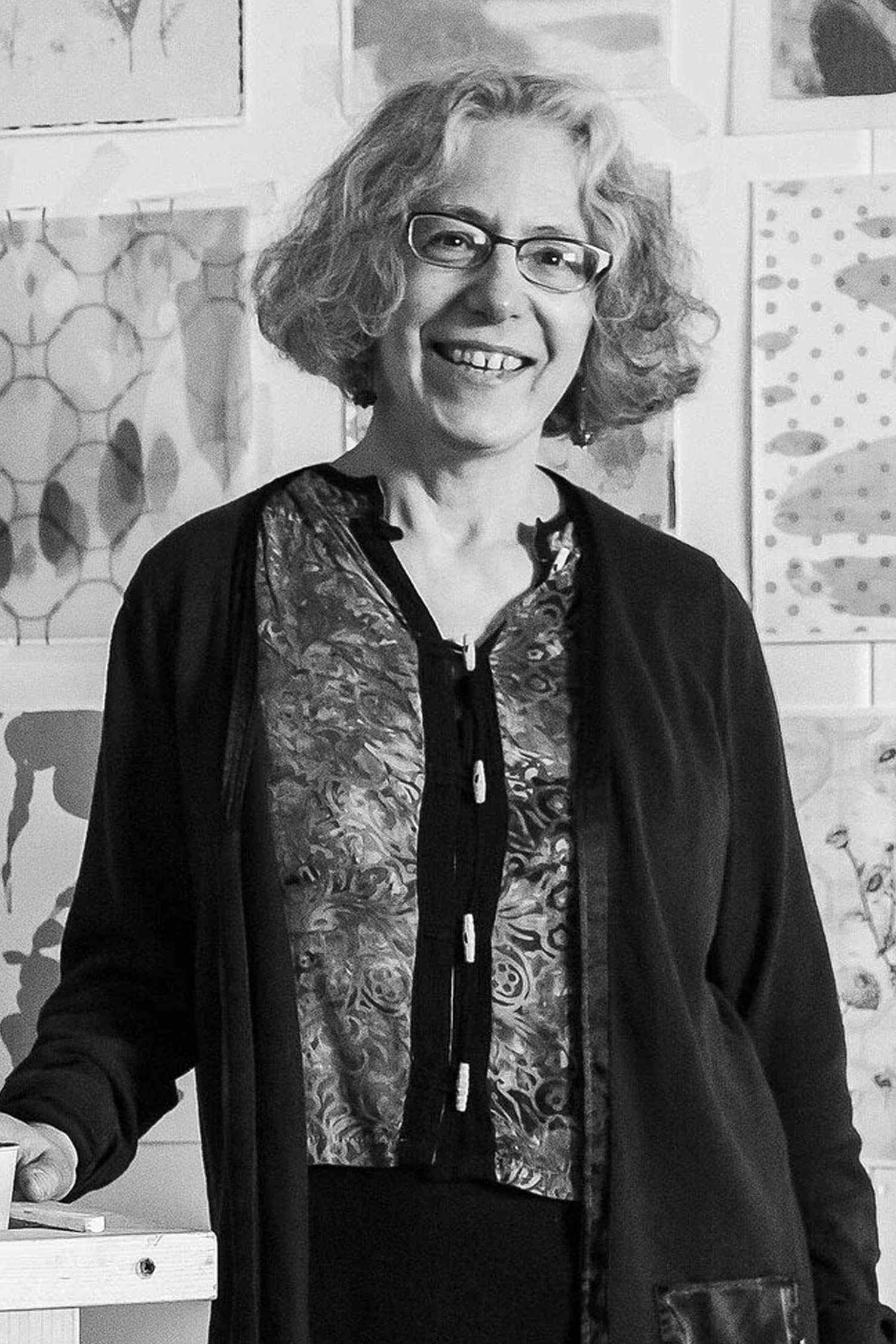 Black-and-white portrait of a smiling woman with short curly hair and glasses standing in front of a wall filled with artwork.