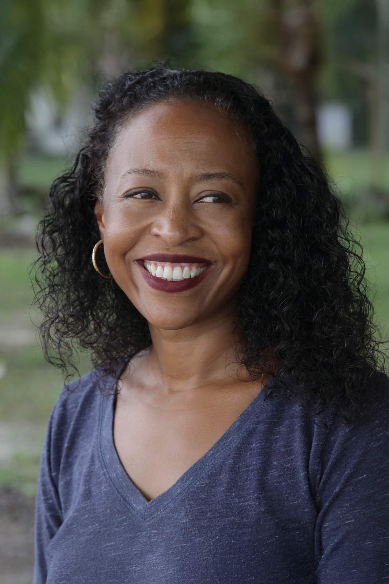 Portrait of Ladee Hubbard smiling outdoors, with long curly hair and a softly blurred green background.