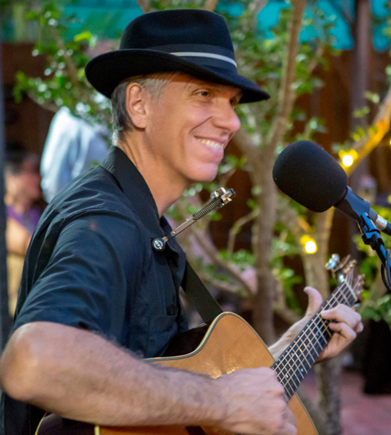 Lance Taylor smiling while playing acoustic guitar at an outdoor performance.