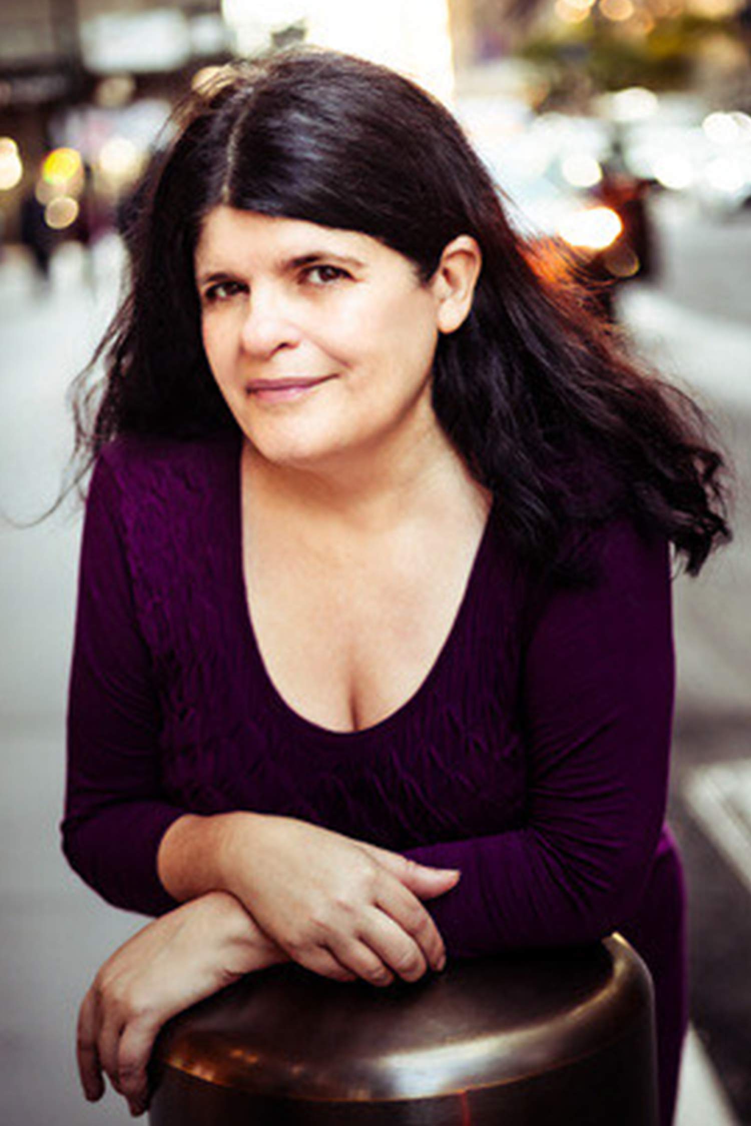 Portrait of a woman with long dark brown hair wearing a deep purple top, leaning with her arms crossed over the back of a chair on a softly blurred city street.