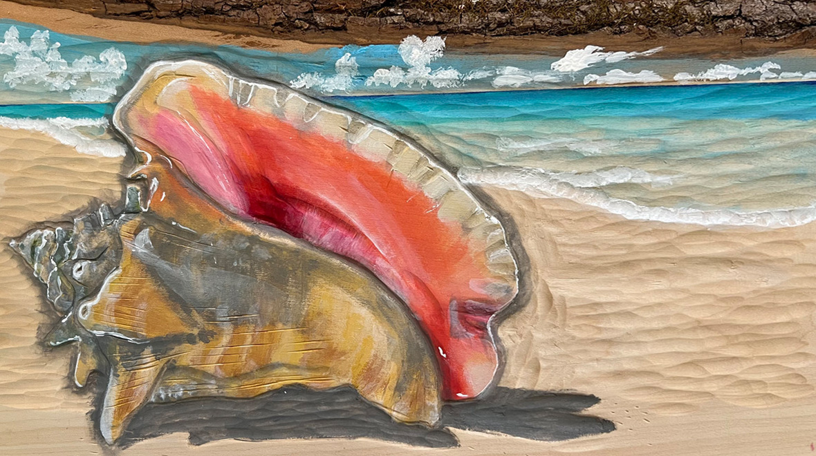 Painted relief carving of a large pink and orange conch shell resting on a sandy beach, with turquoise water, breaking waves, and clouds in the background.