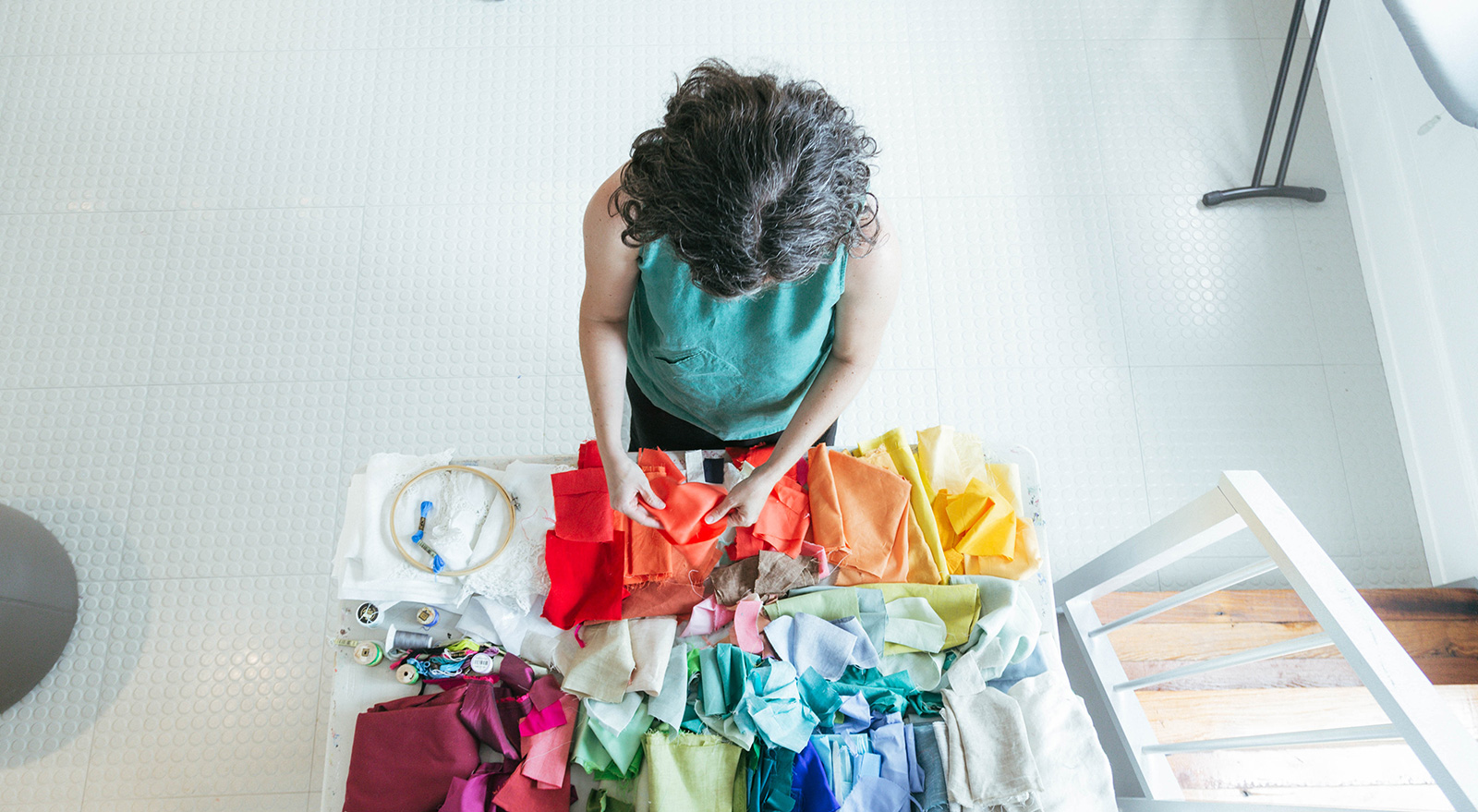 Artist Lee Jensen works at a table covered with fabric scraps arranged by color, alongside embroidery supplies.