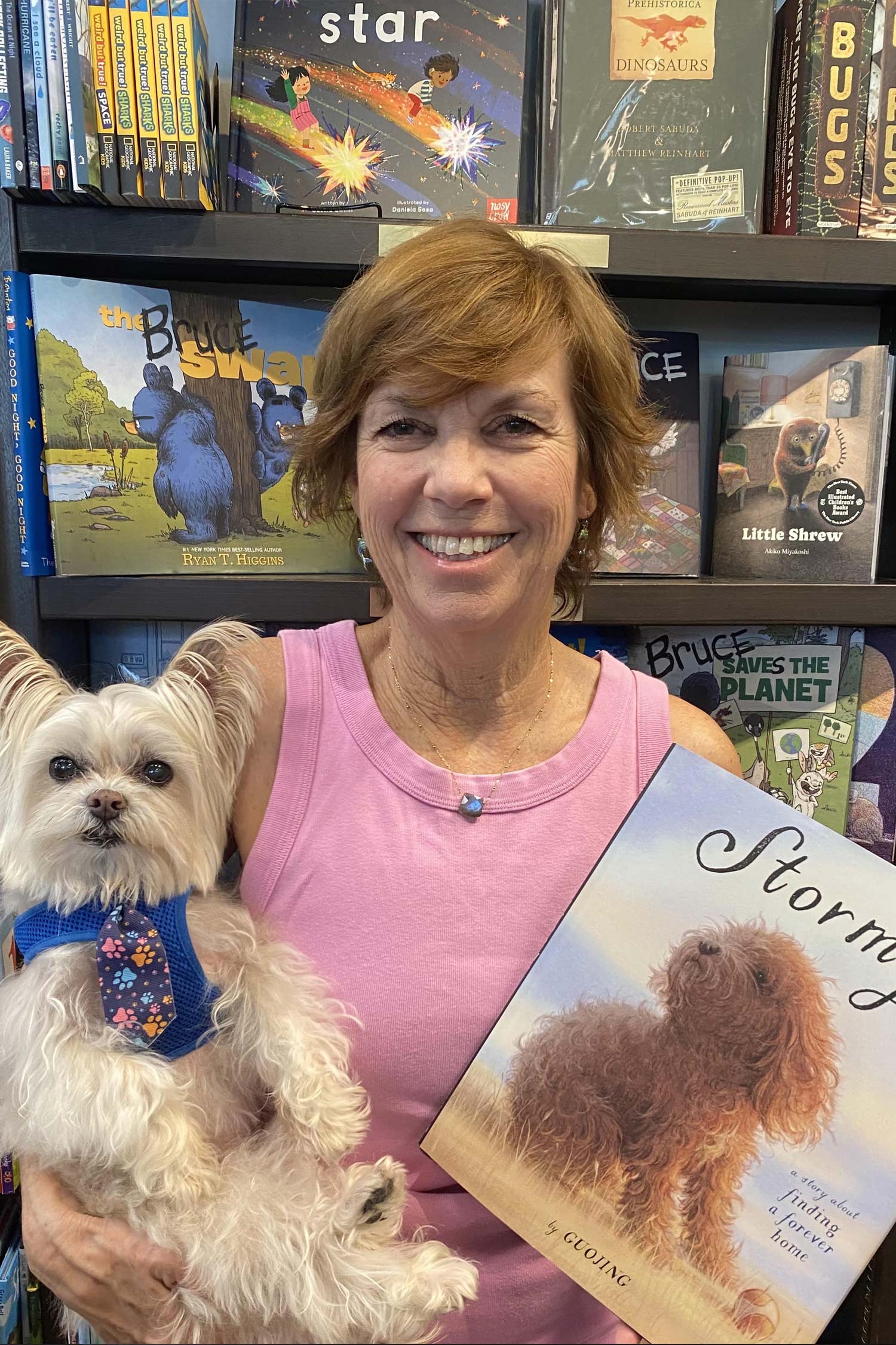 Smiling woman in a pink sleeveless top holds a small white dog and a children’s book while standing in front of bookstore shelves.