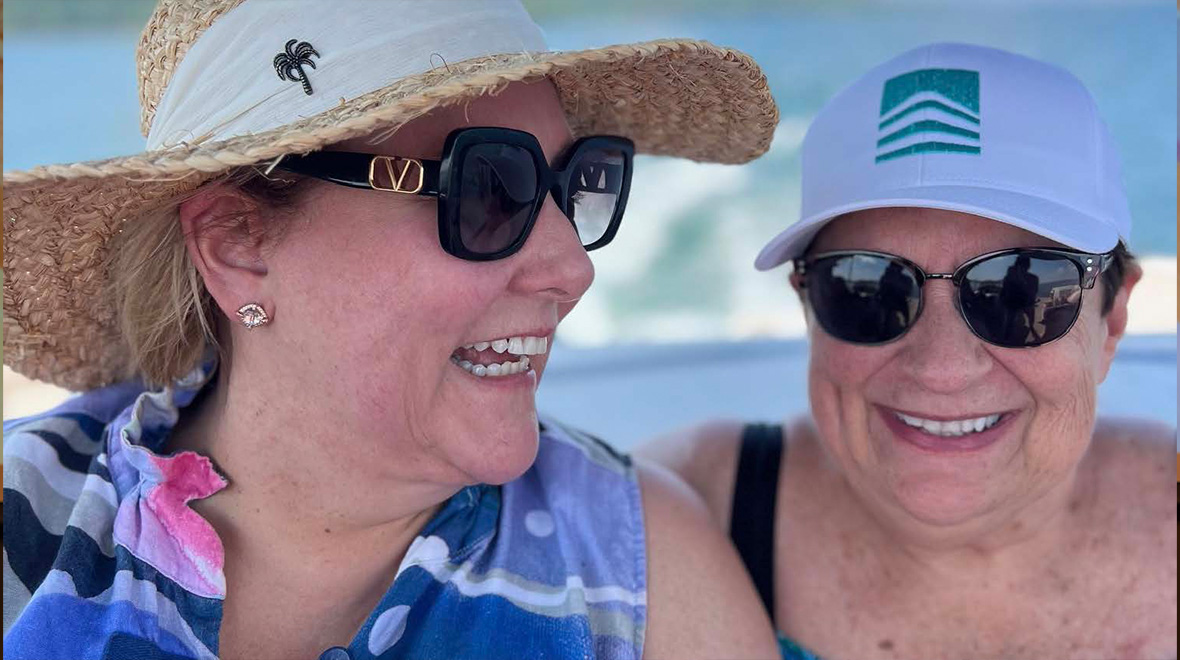 Two women wearing sun hats and sunglasses smile together outdoors.