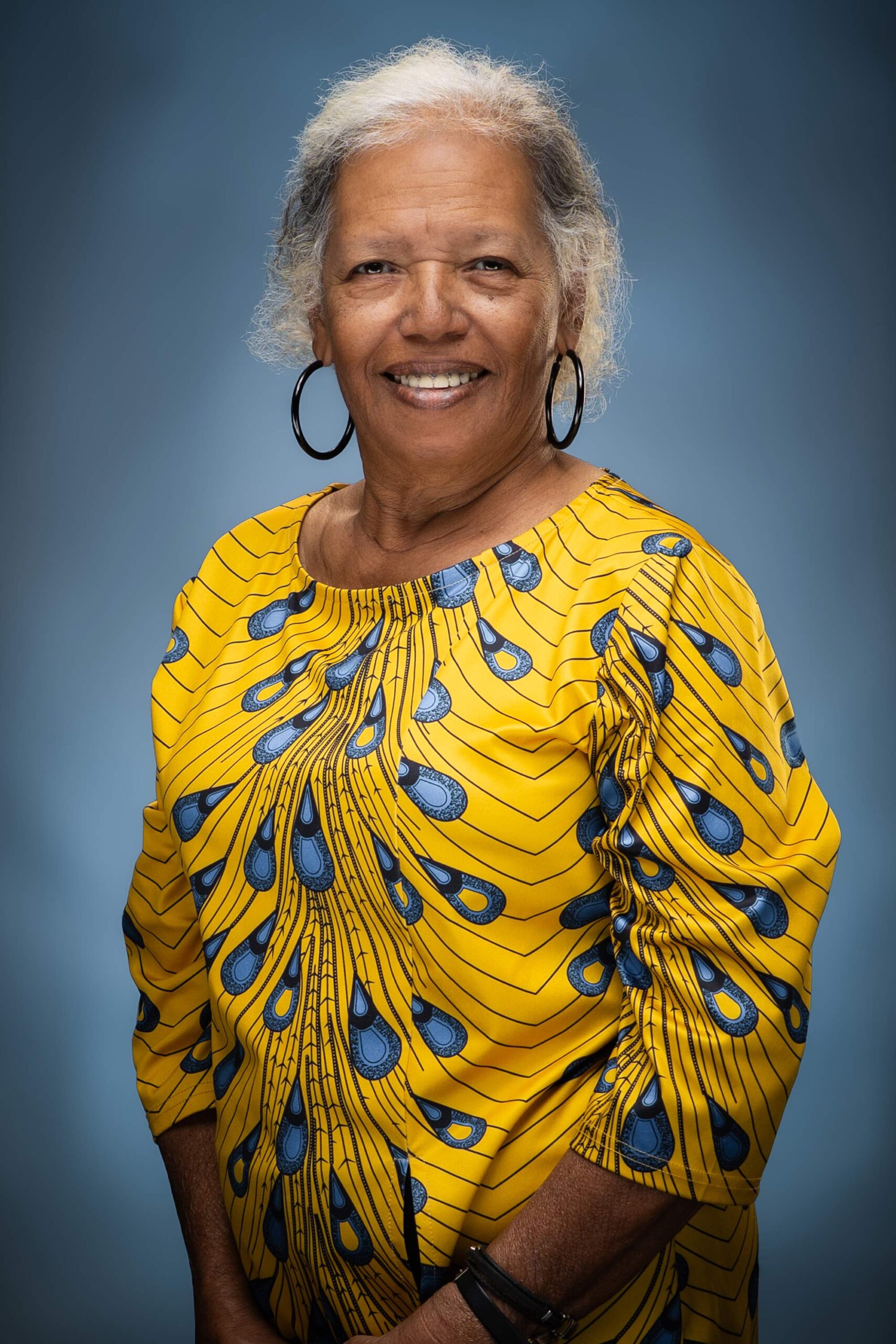 Portrait of Lori smiling in a vibrant yellow and blue patterned top with hoop earrings against a blue studio background.