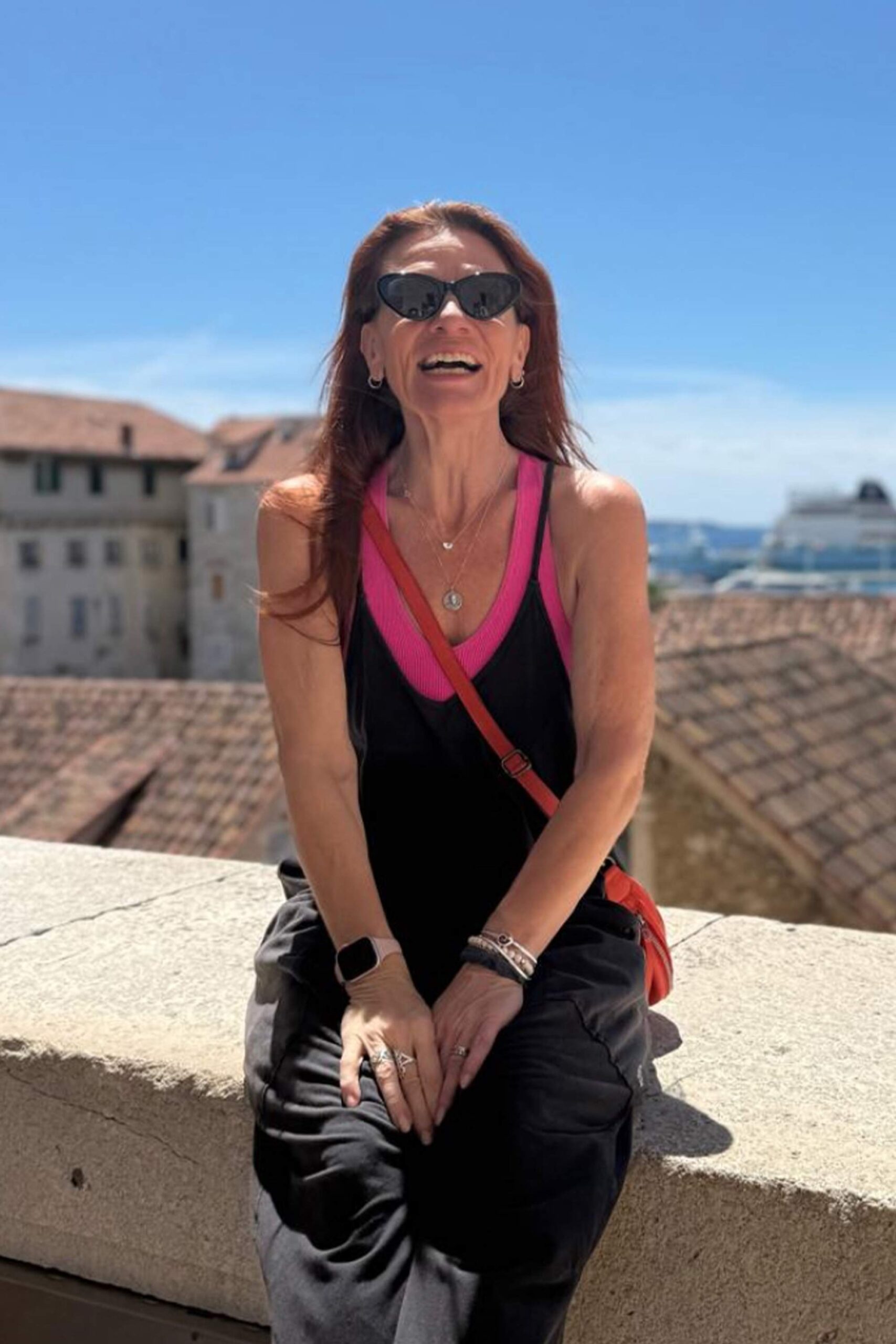 Jacqueline Luhta smiling outdoors in sunglasses, with sunlit rooftops and blue sky in the background.