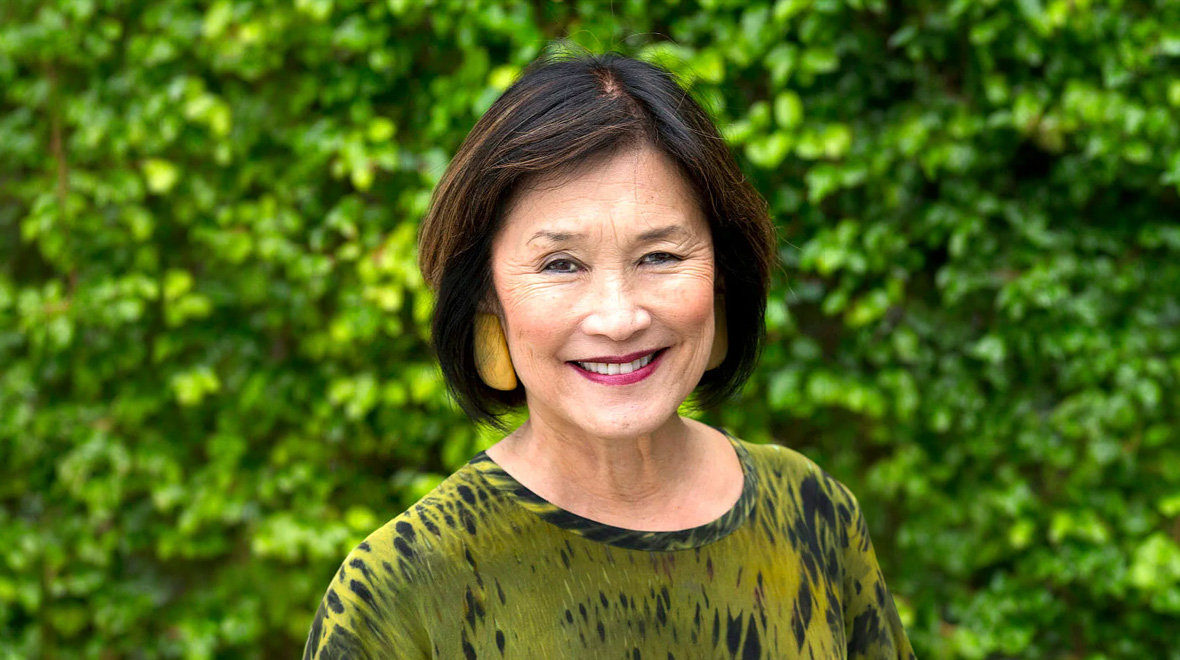 Smiling woman with short dark hair and yellow earrings, photographed outdoors against lush green foliage.
