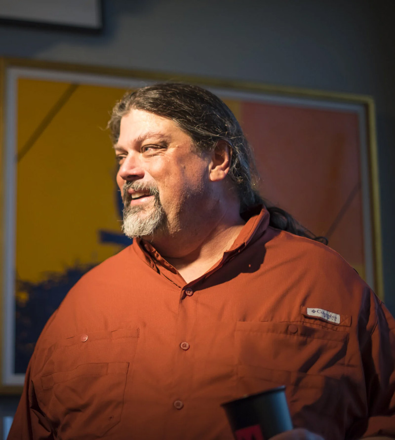 Mark Hedden smiling, with a brightly colored abstract artwork behind him.