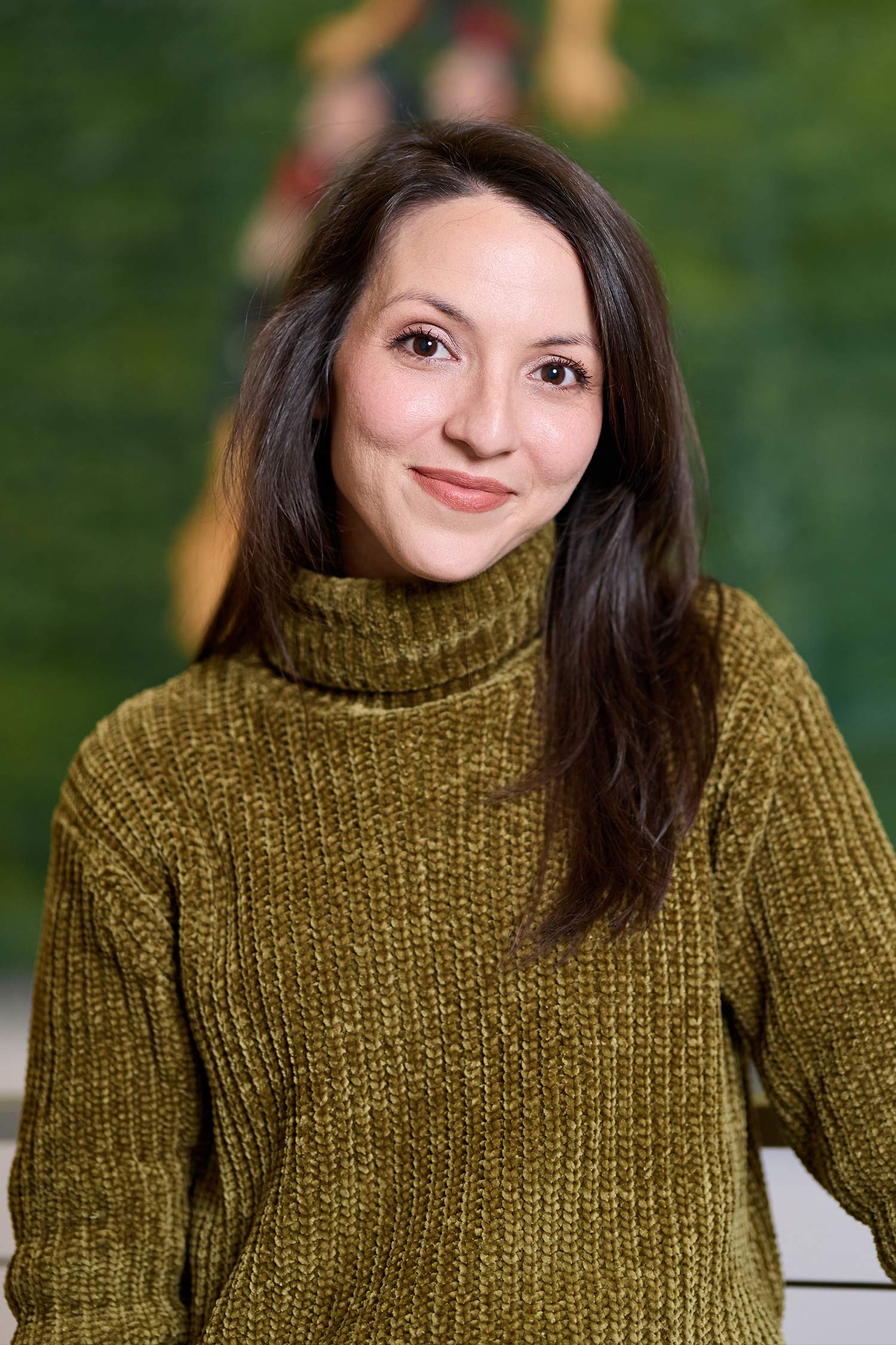 Portrait of a smiling woman with long dark brown hair wearing an olive green turtleneck sweater, posed against a softly blurred green background.