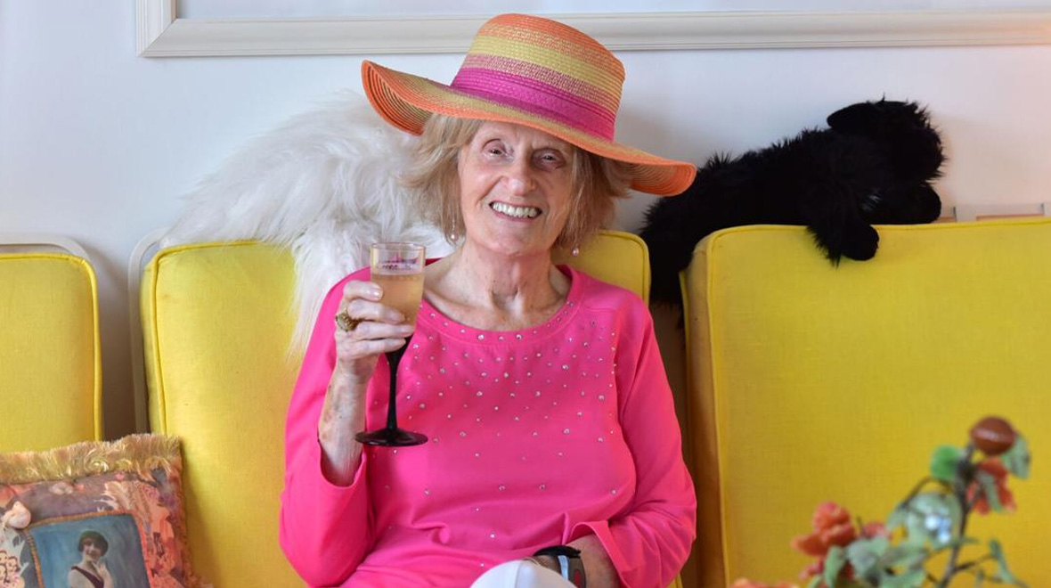 Smiling older woman in a bright pink top and wide-brimmed striped hat raises a glass while seated on a yellow sofa.