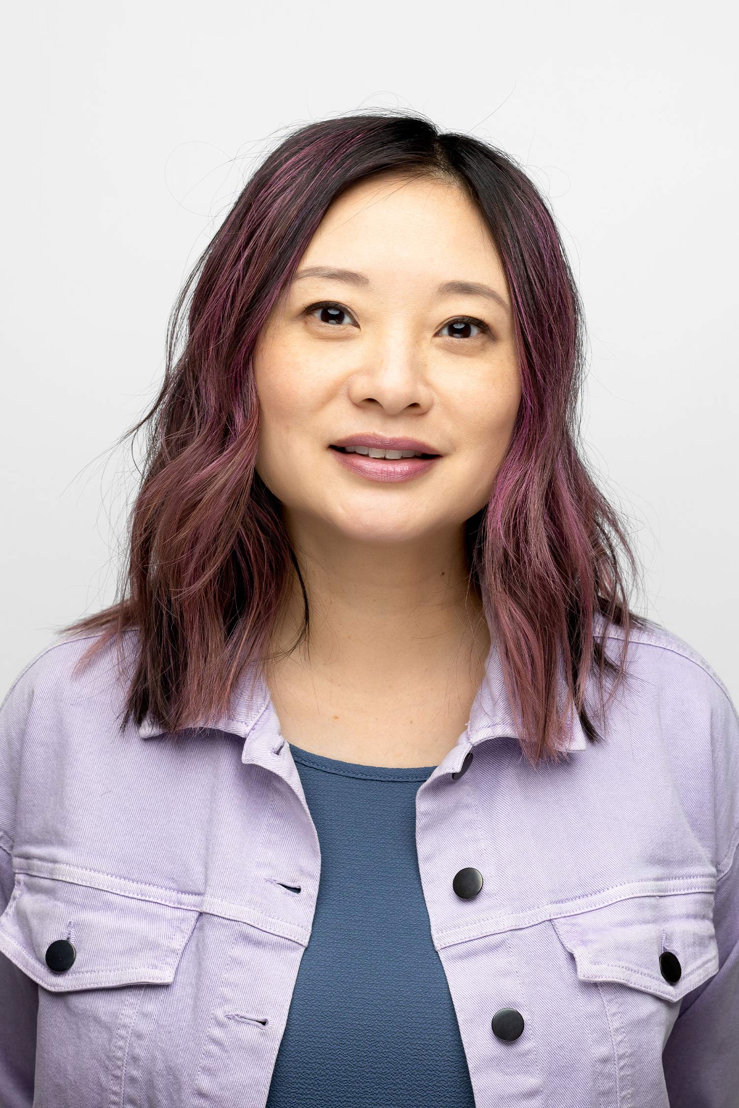 Head-and-shoulders portrait of a woman with shoulder-length dark hair tinted purple, wearing a pale lavender jacket over a blue-gray top against a plain white background.