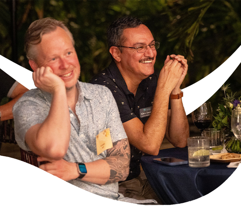 Two men smile and laugh while seated at a table during an outdoor evening gathering at The Studios of Key West.