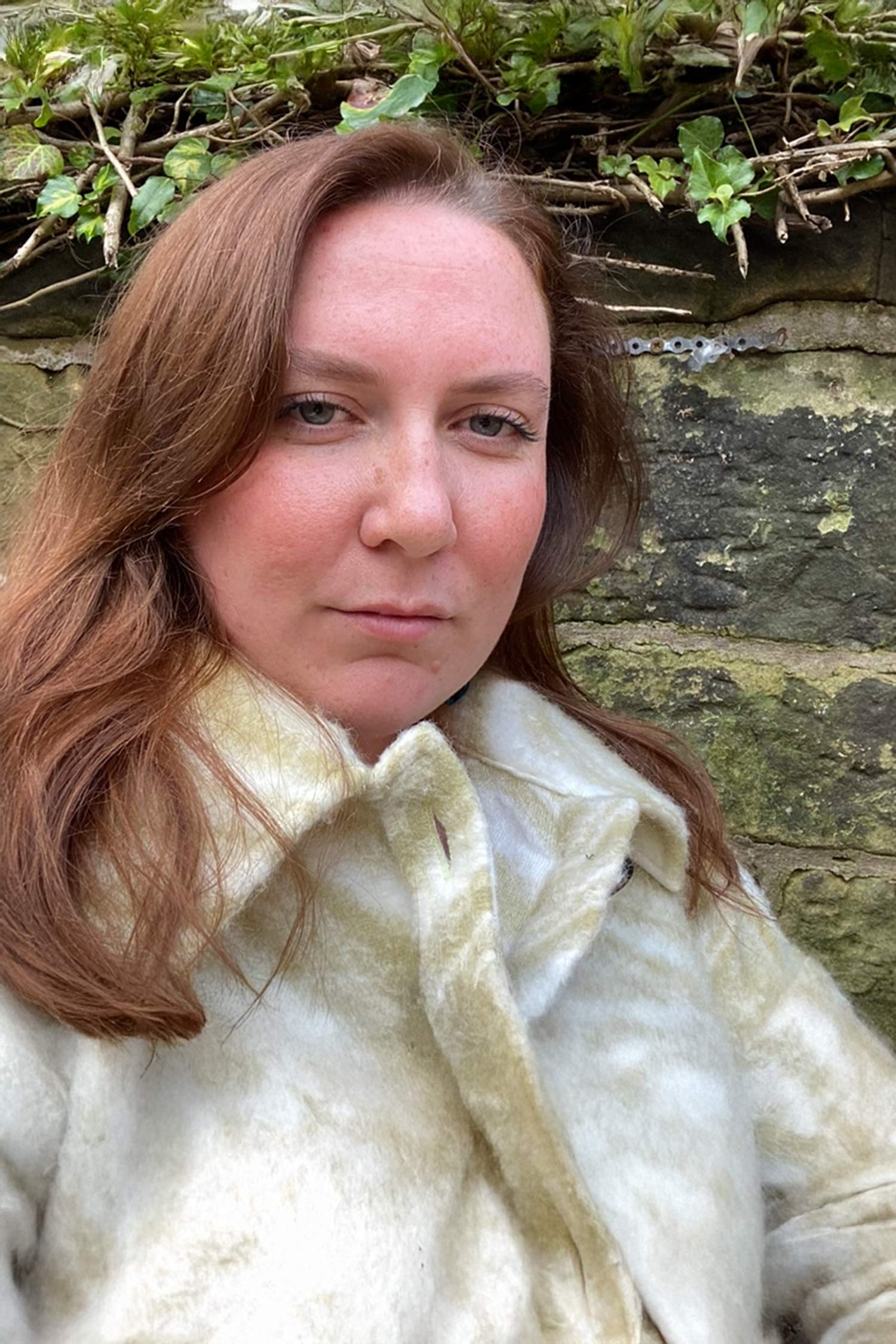 Portrait of Meredith MacLeod Davidson with long auburn hair, wearing a cream-colored coat in front of a stone wall and greenery.