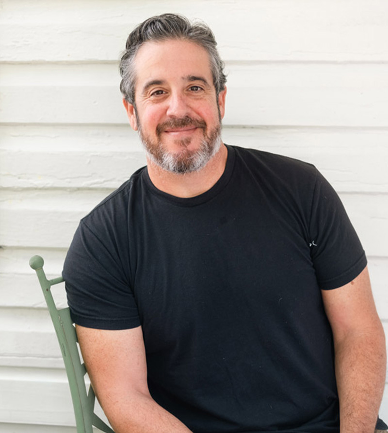 Mike Marrero smiling while seated against a white wooden wall.