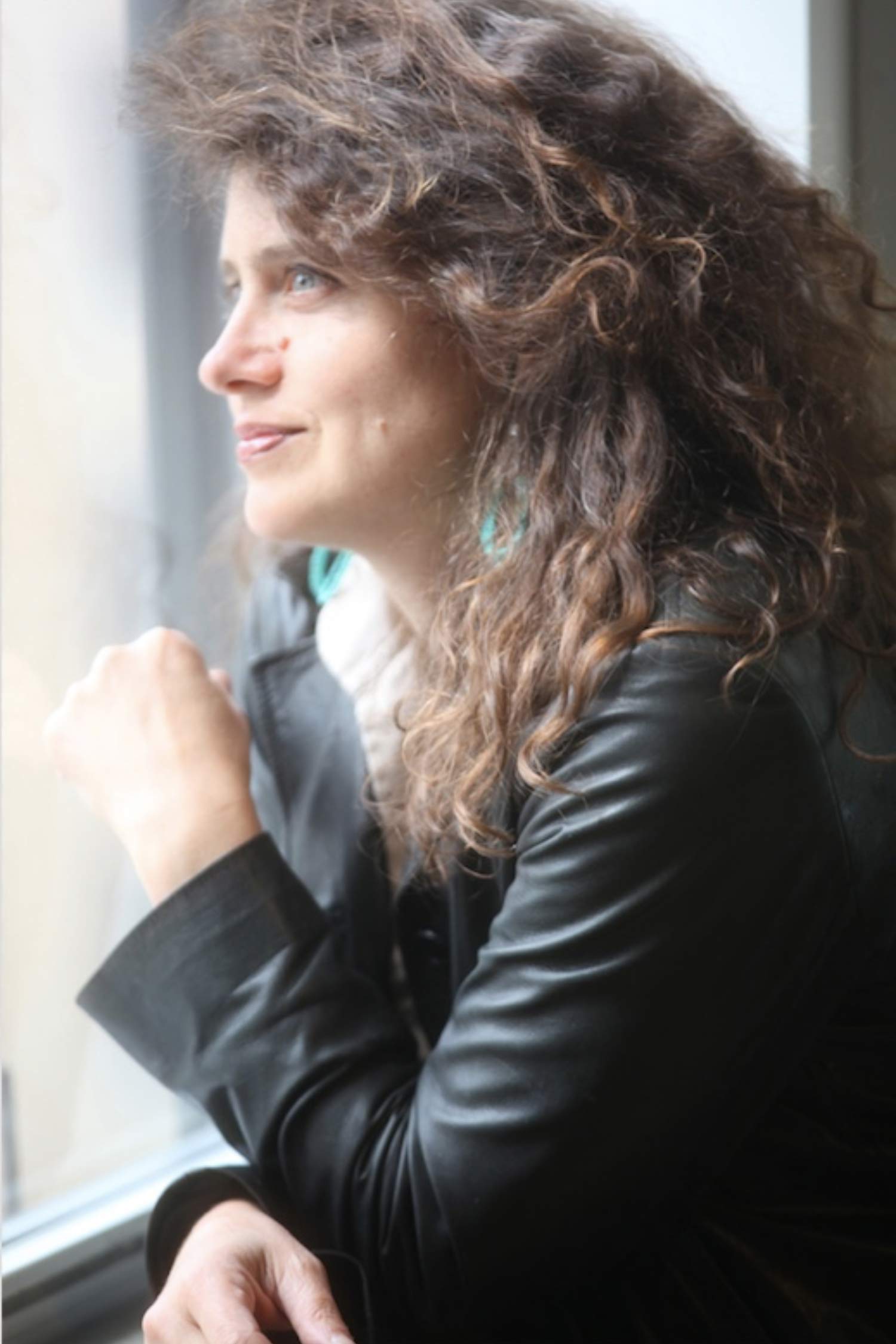 Side-profile portrait of Nell Breyer seated by a window, looking outside with curly brown hair and wearing a black jacket.