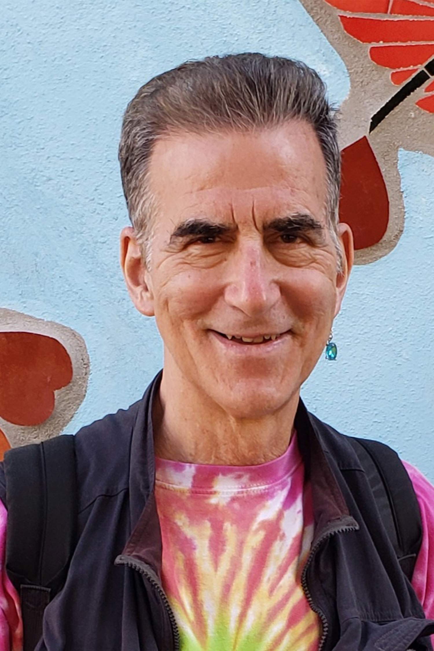 Portrait of Rich Orloff smiling in a colorful tie-dye shirt and dark vest against a painted blue wall.