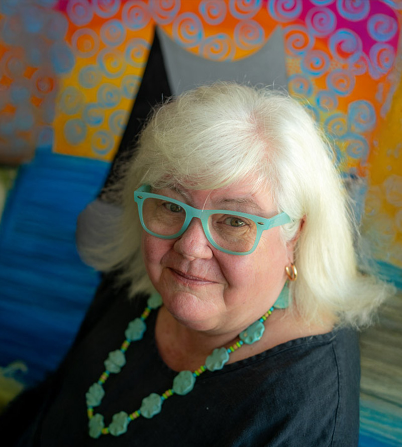 Pam Hobbs wearing turquoise glasses and jewelry, with a colorful painting behind her.