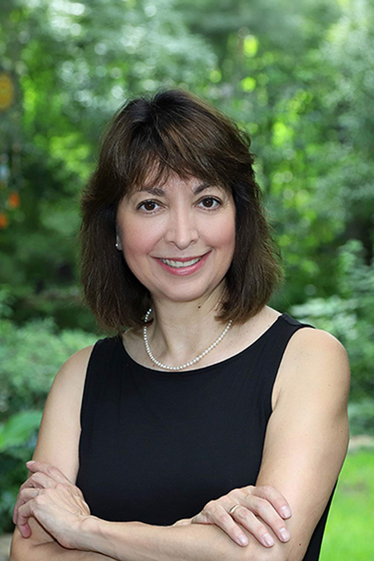 Pamela Anne Quiroz smiles at the camera with her arms crossed, standing outdoors against a leafy green background.