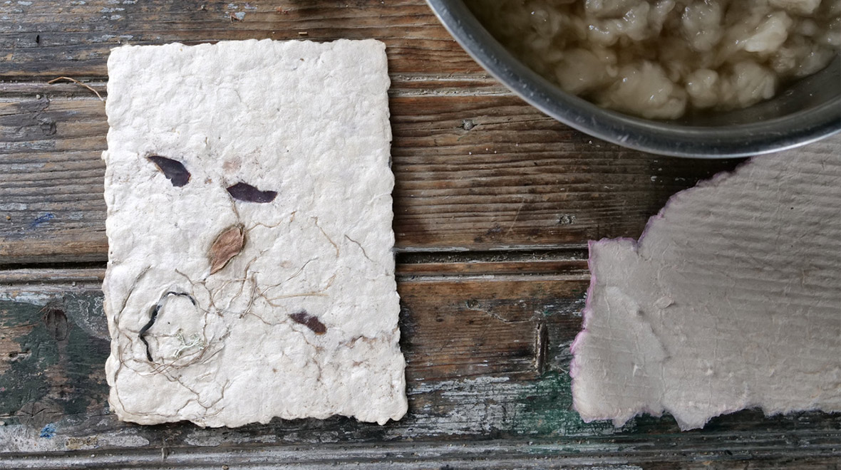 Handmade paper with embedded leaves and plant fibers beside a bowl of wet paper pulp on a rustic wooden worktable.