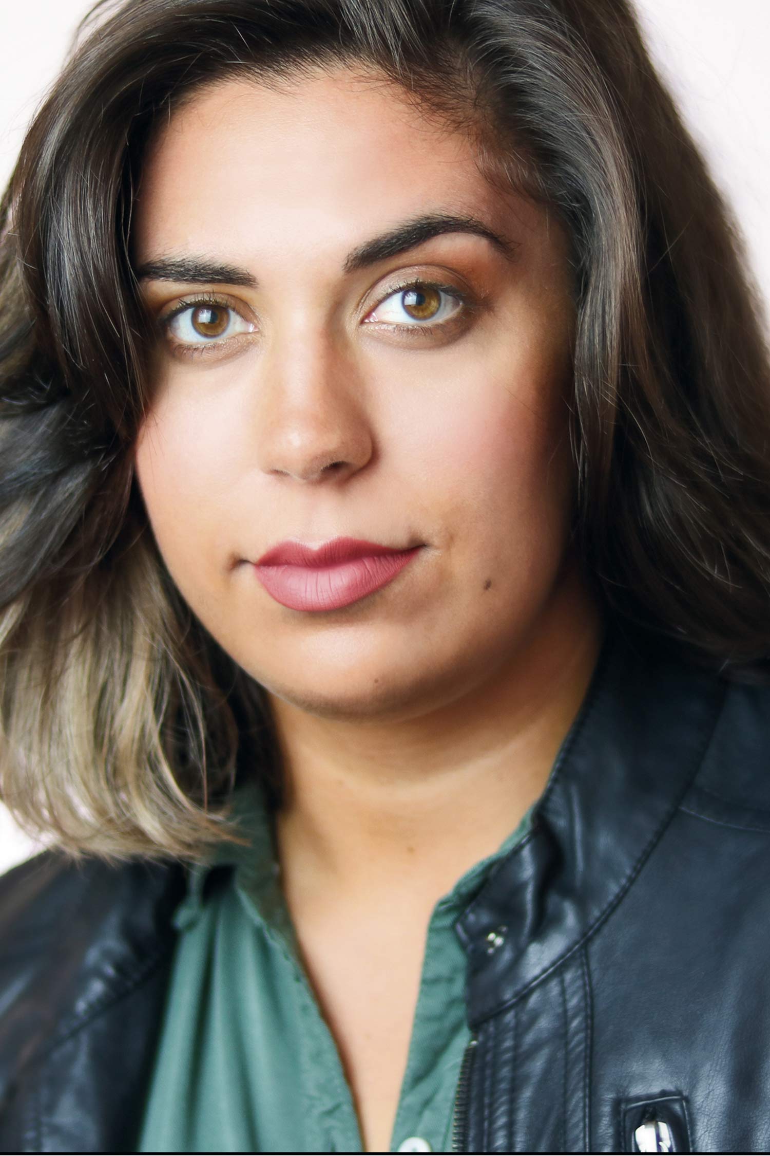 lose-up headshot of a woman with shoulder-length dark brown hair with lighter ends, brown eyes, and a neutral expression, wearing a green blouse and black leather jacket against a light background.