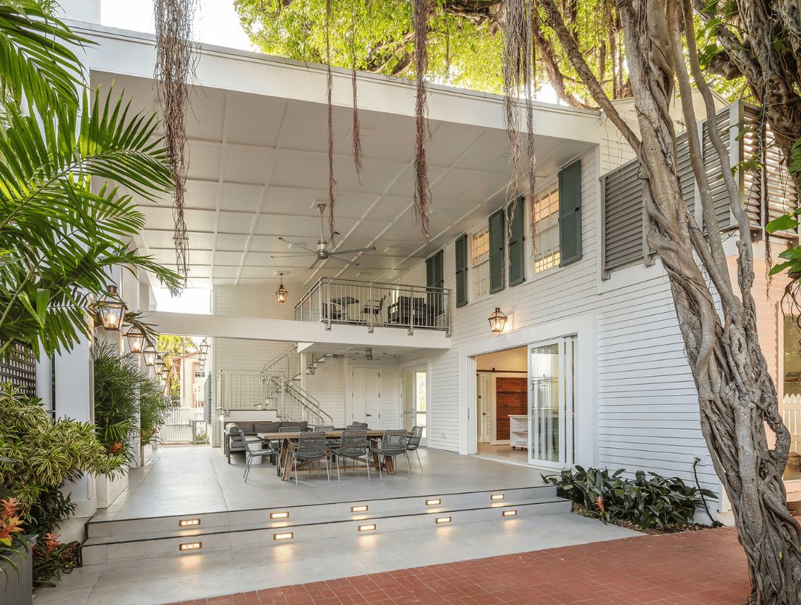 A covered outdoor courtyard of a white two-story house, with a long dining table and chairs on a raised concrete patio, framed by a large banyan tree and tropical palms.
