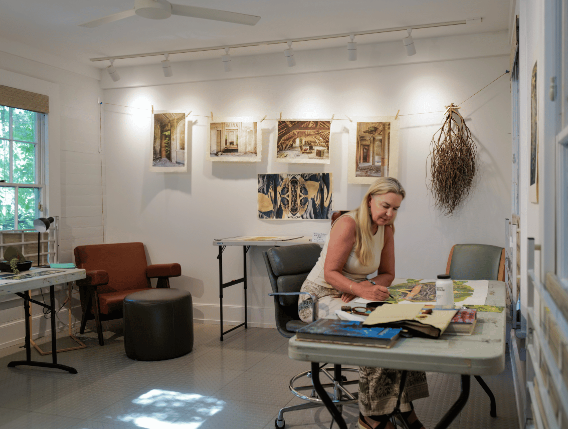 An artist works at a table in a bright white studio at the PEAR House, with a line of paintings clipped to a wire on the wall behind her and a hanging bundle of dried roots to one side.