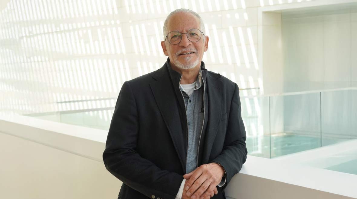 An older man with glasses and a gray beard, wearing a dark jacket, stands in a bright modern interior with geometric patterns of sunlight behind him.