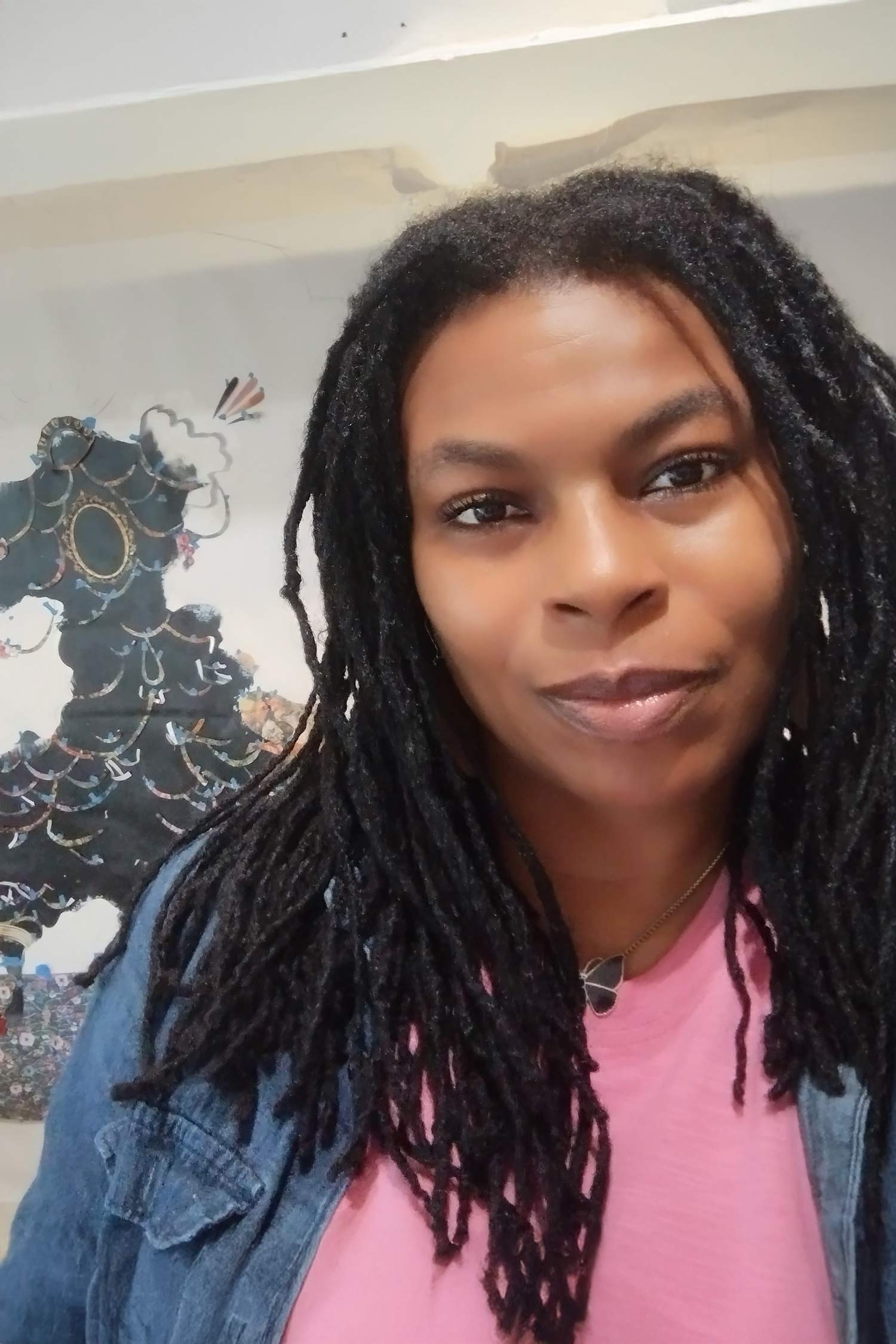 Portrait of a person with long locs wearing a pink shirt and denim jacket, photographed in front of artwork.