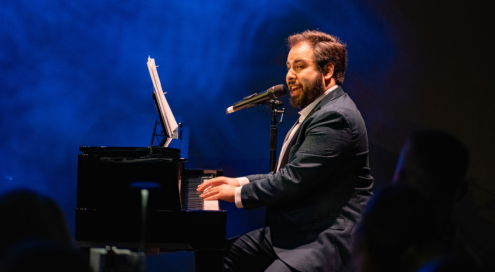 A pianist sings into a microphone while playing a grand piano onstage under dramatic blue lighting.