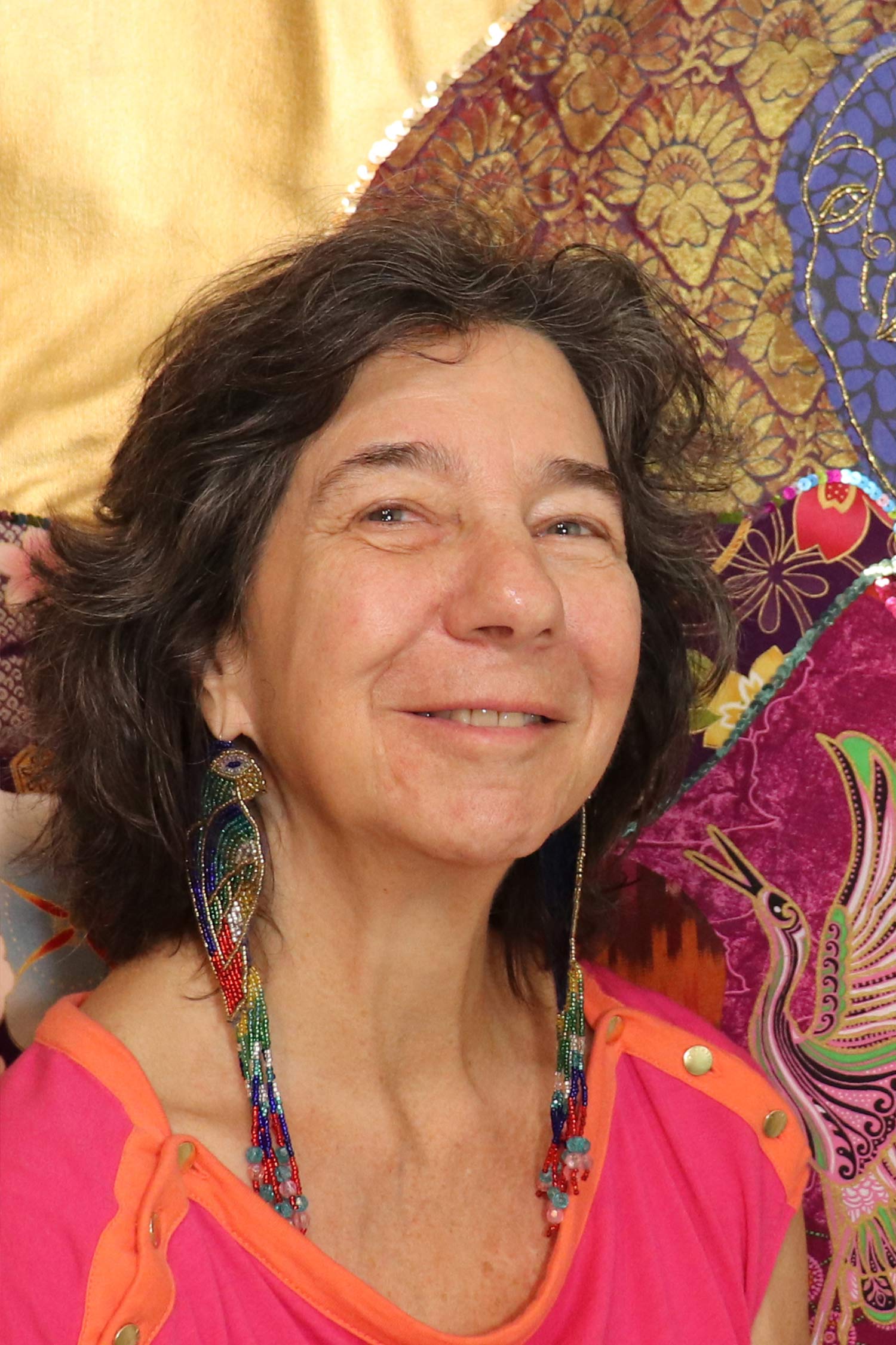 Close-up portrait of a smiling woman with short dark hair, wearing long multicolored beaded earrings and a bright pink and orange top, with richly patterned textile artwork behind her.